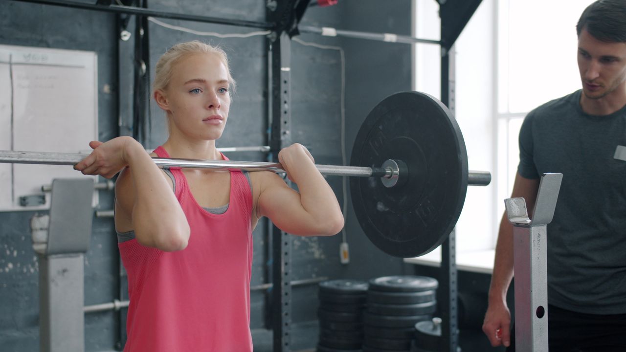 Young woman practicing weightlifting with trainer's guidance in gym.