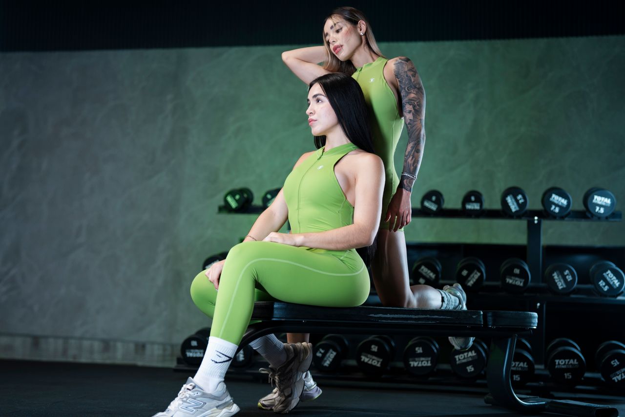 Two women in green activewear posing confidently on a gym bench surrounded by weights.